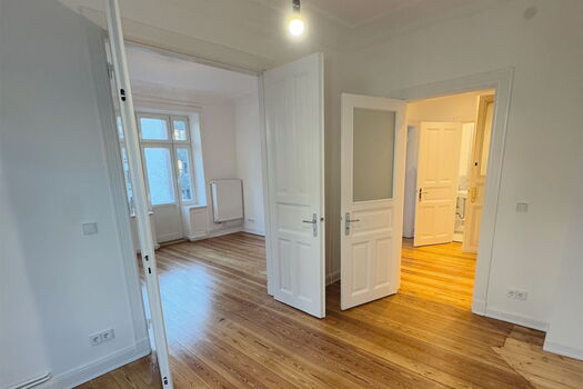 Ein heller, leerer Raum mit Holzfußboden, weißen Wänden und offenen weißen Türen, die zu anderen Räumen führen. Große Fenster lassen natürliches Licht herein, und unter einem Fenster ist ein Heizkörper zu sehen.