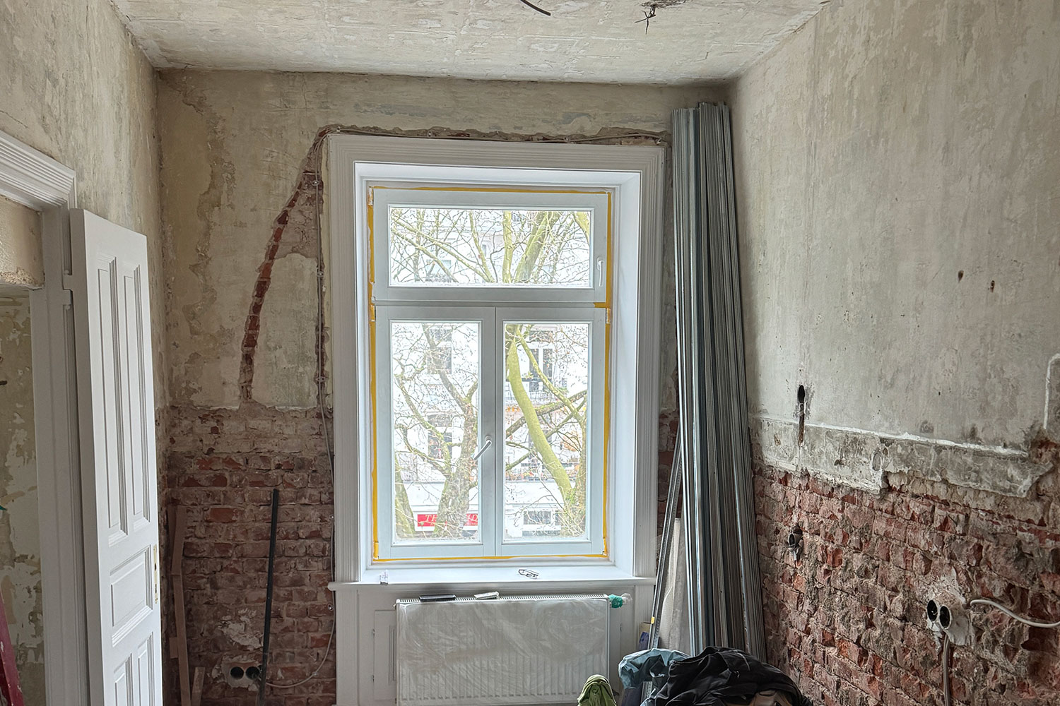 Ein kleiner Raum, der gerade renoviert wird, mit freiliegenden Ziegelwänden, teilweise abgetragenem Putz, einem weißen Fenster mit einem Heizkörper darunter und verstreutem Renovierungsmaterial. Vor dem Fenster ist ein Baum zu sehen.