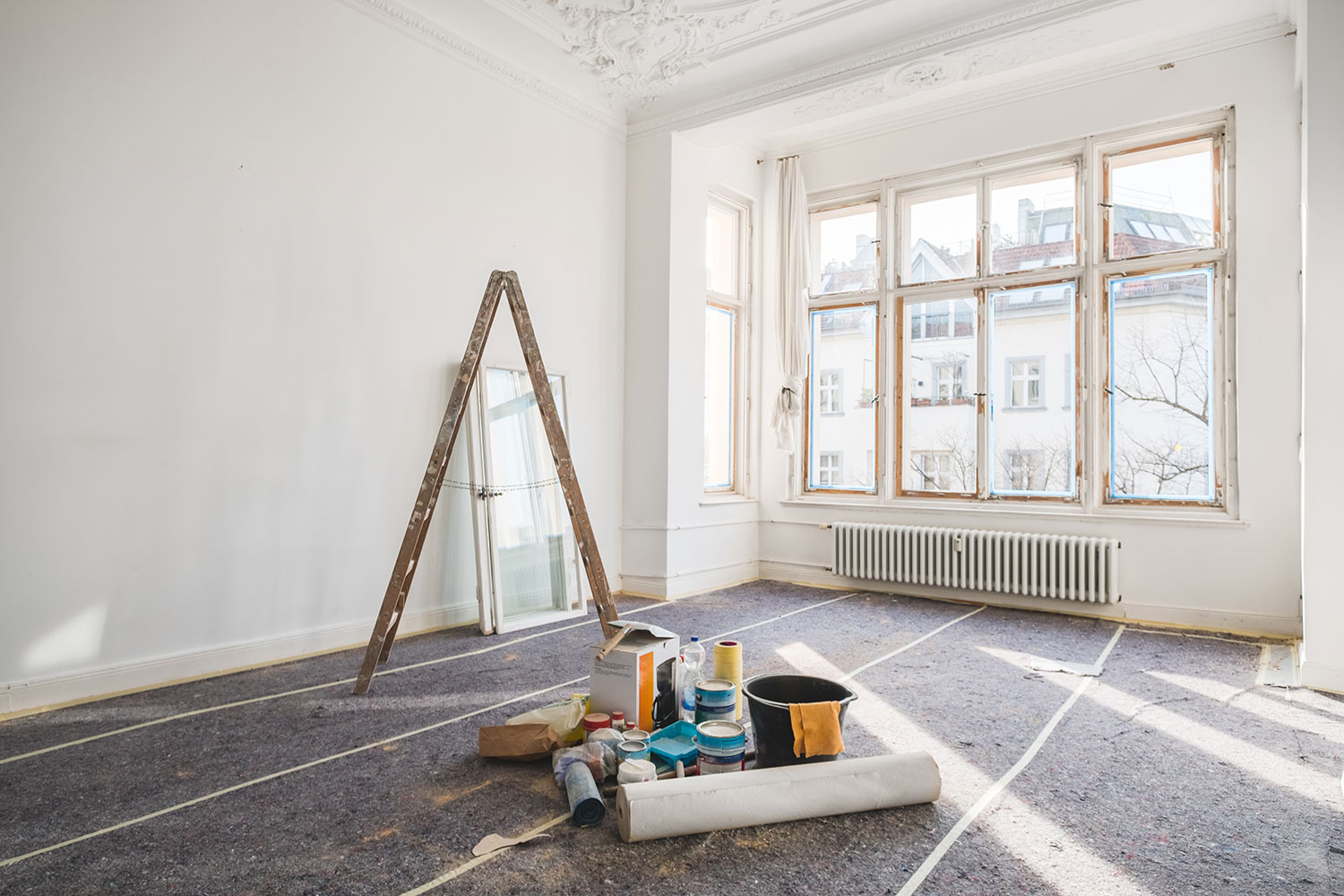 Ein heller, leerer Raum mit großen Fenstern, einer Trittleiter, einem Spiegel und auf dem Boden verstreuten Malutensilien, die auf eine laufende Renovierung hindeuten. Sonnenlicht strömt herein und erhellt den unfertigen Raum.